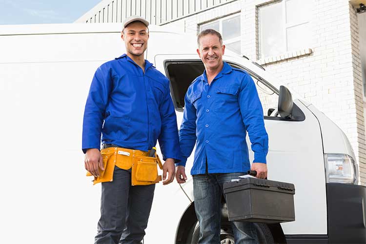 Two smiling locksmiths in blue uniforms standing next to a white van; one holding a toolbox and the other with a tool belt.